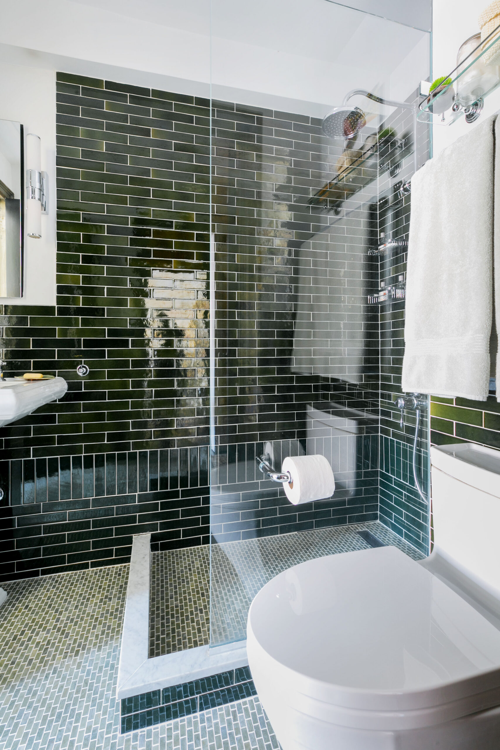 Gramercy bathroom with a glass-mounted toilet paper holder, clear shower panel, glossy green wall tile, chrome fixtures, and mosaic flooring.