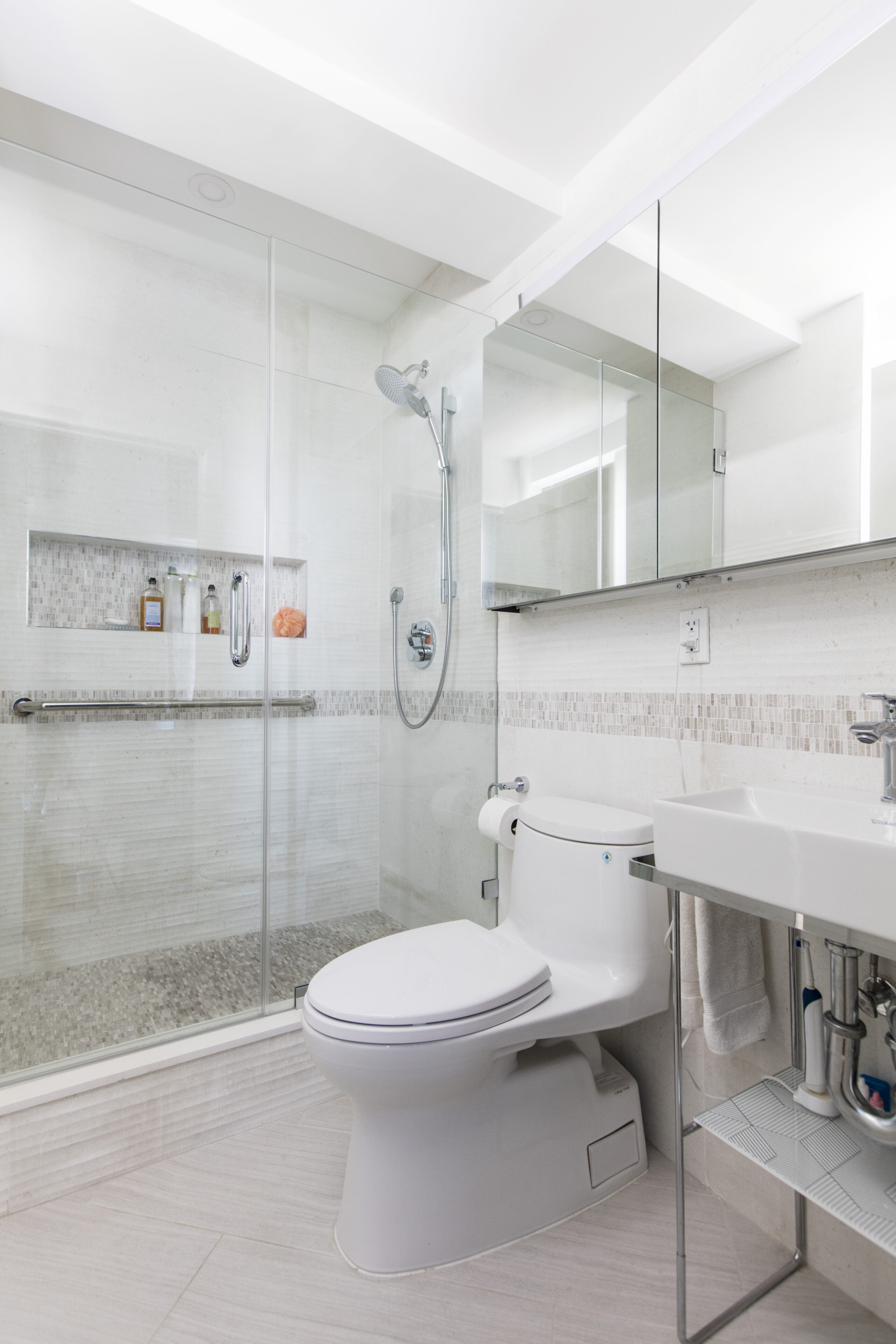 Kensington bathroom with a glass-enclosed shower, long grab bar, handheld shower, wall-mounted sink, mirrored cabinet, and pale tile.