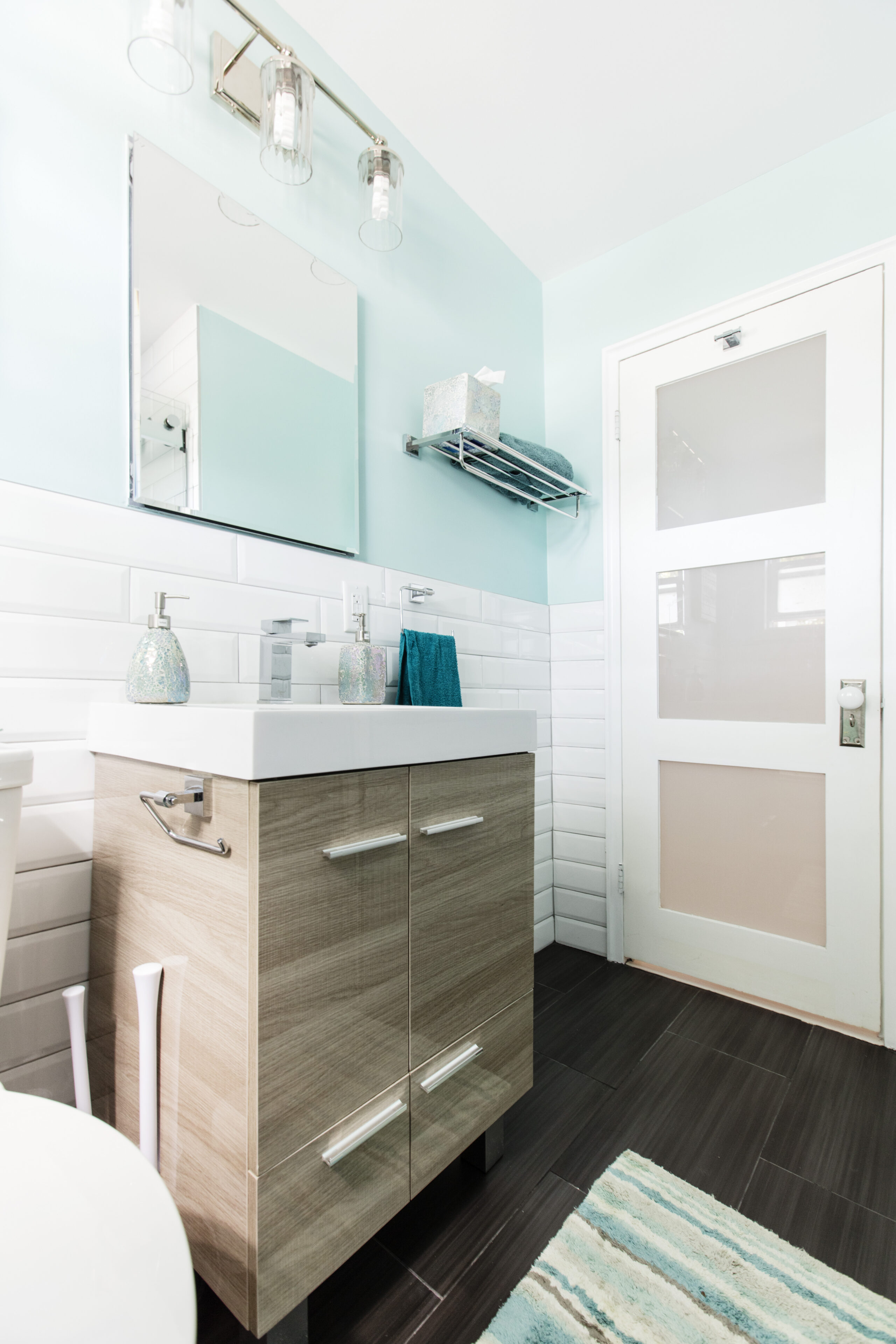 Long Island bathroom with a chrome toilet paper holder mounted to the vanity, long cabinet pulls, a train rack, white subway tile, and aqua walls.