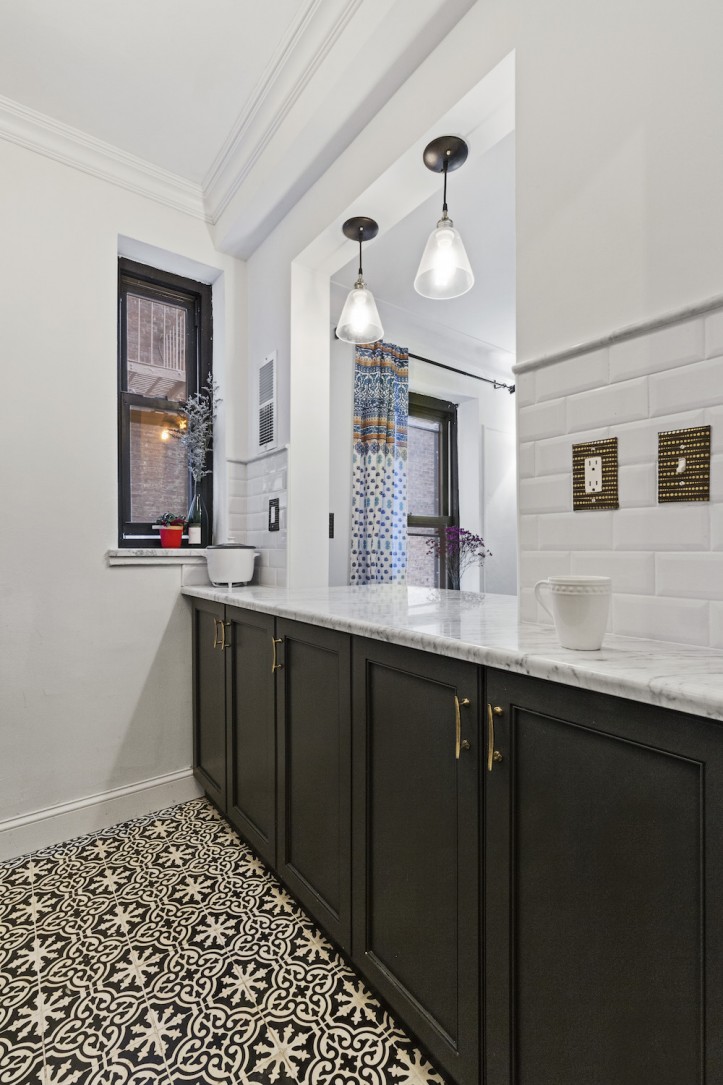 Manhattan galley kitchen with a pass-through opening, charcoal cabinets, brass pulls, marble counters, white subway tile, and patterned flooring.