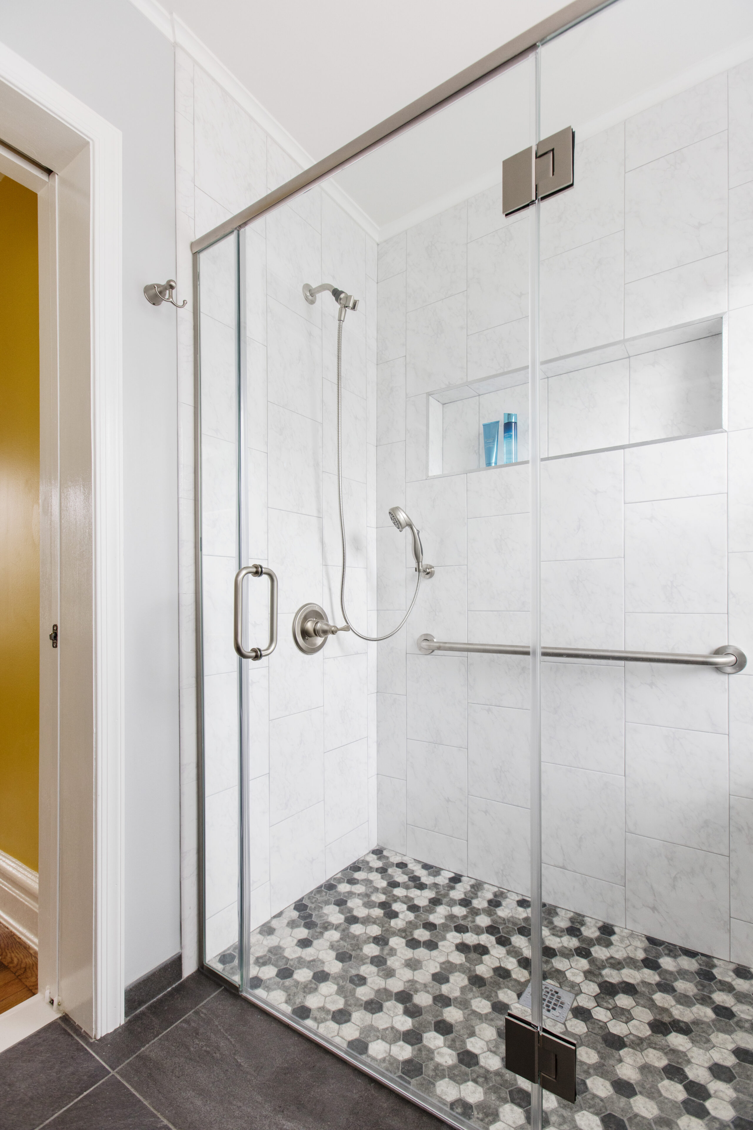 Queens bathroom with a glass walk-in shower, long brushed metal grab bar, handheld shower, built-in niche, and black-and-white hex floor tile.