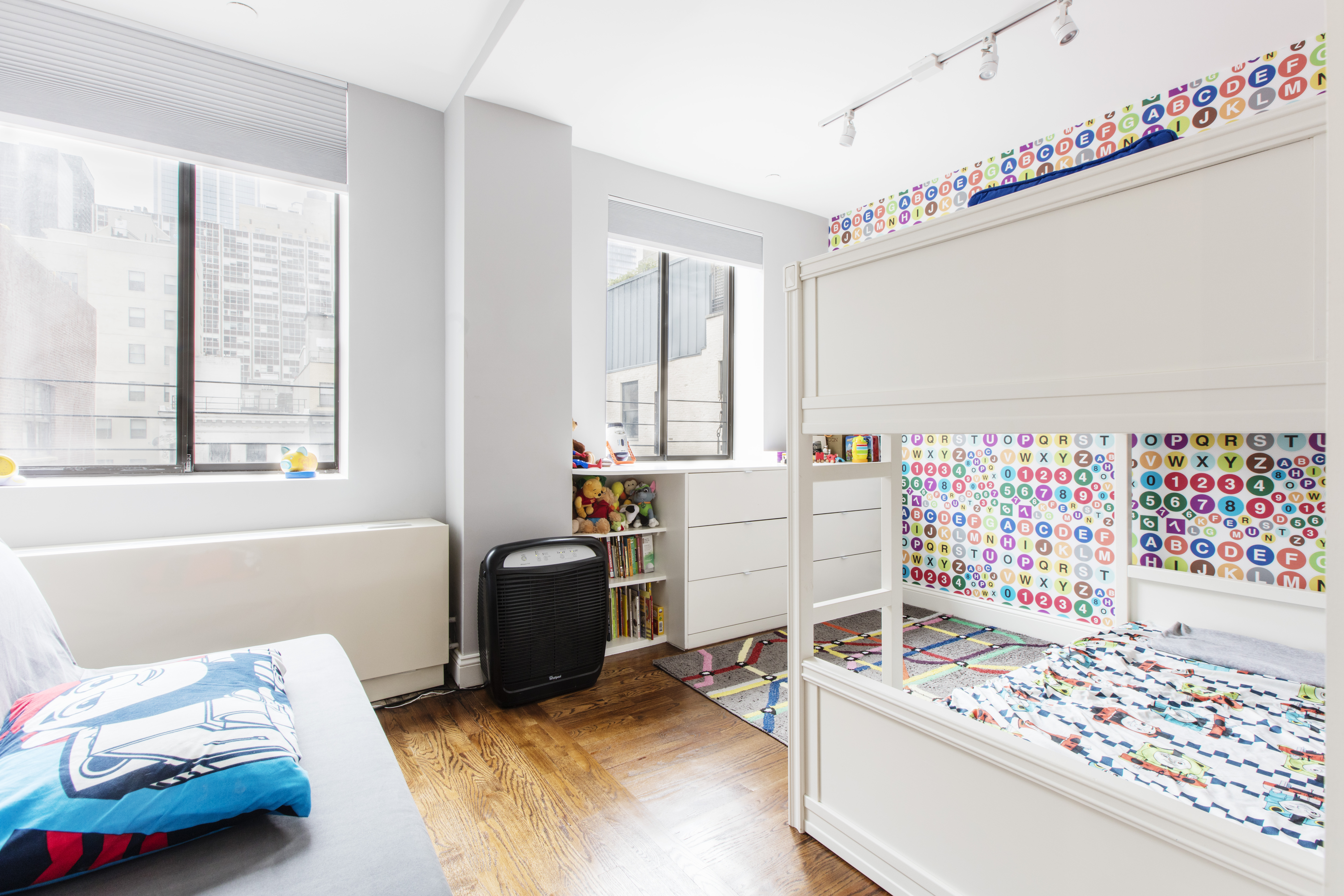 Tribeca extra bedroom idea with white bunk beds, alphabet wallpaper, wide windows, wood floors, built-in shelving, and track lighting.
