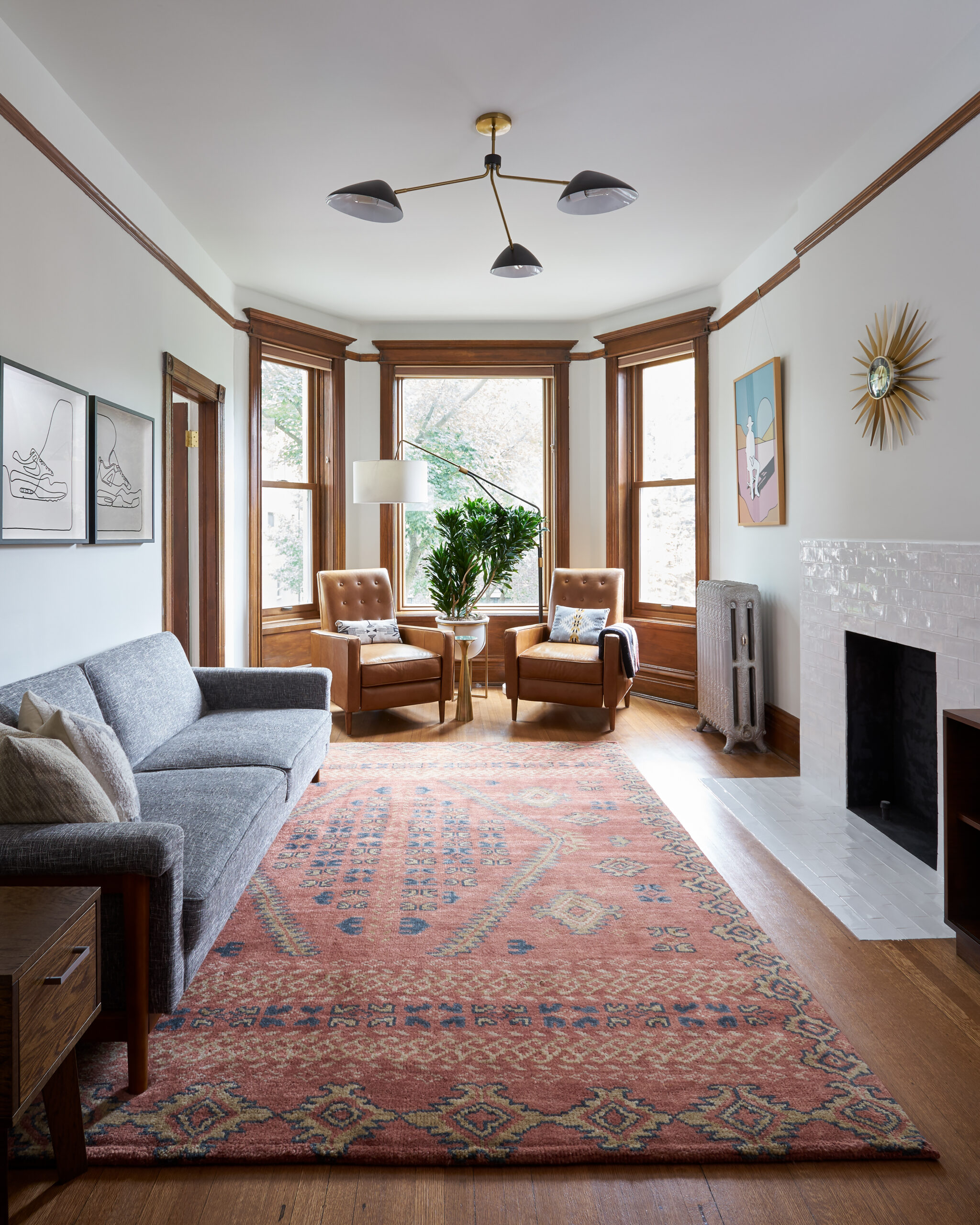 Chicago living room with glossy white subway fireplace tile, original wood window trim, hardwood floors, a patterned rug, and midcentury-style lighting.