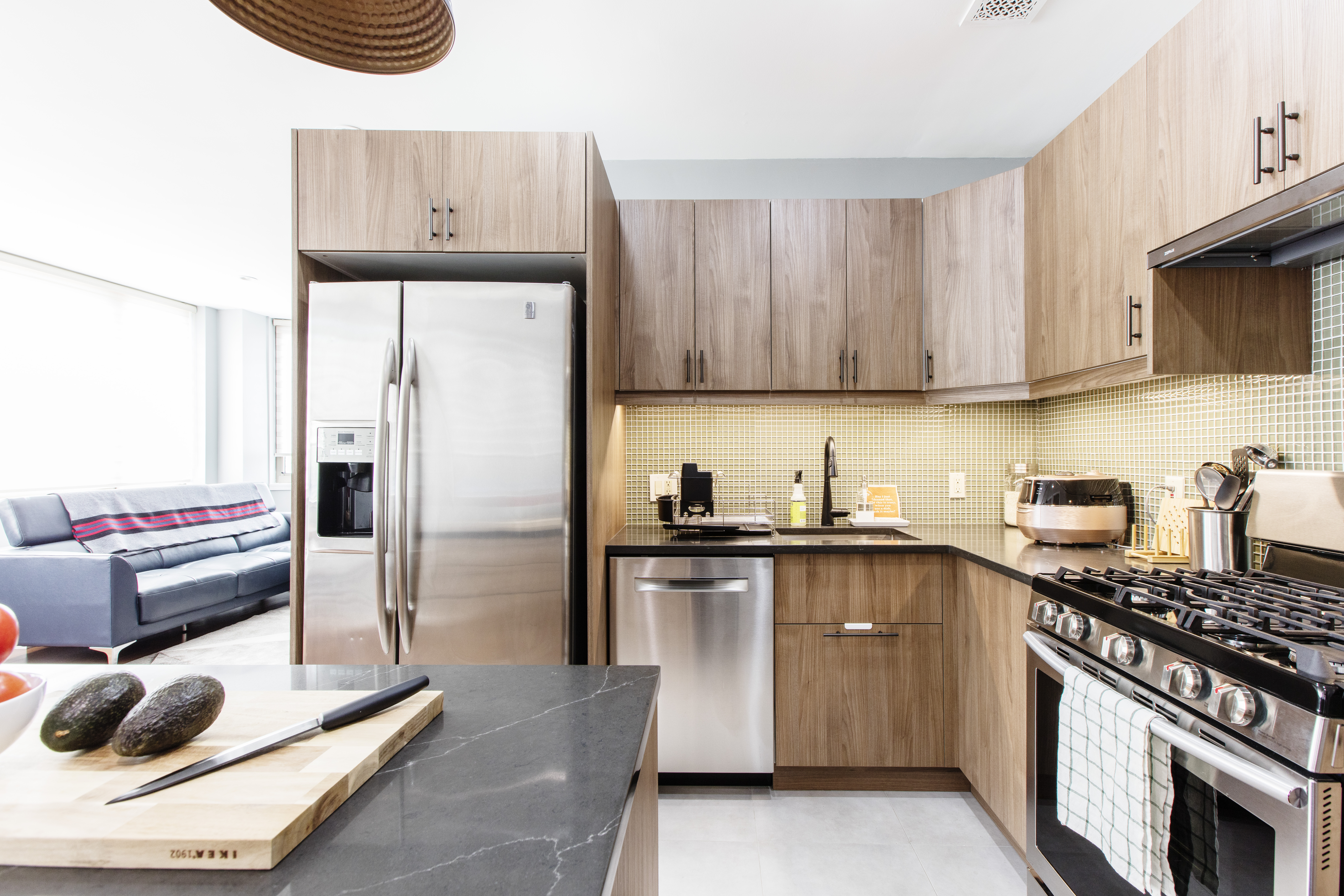 Flushing kitchen with wood corner cabinets, black countertops, mosaic tile backsplash, stainless appliances, and a peninsula beside the living area.