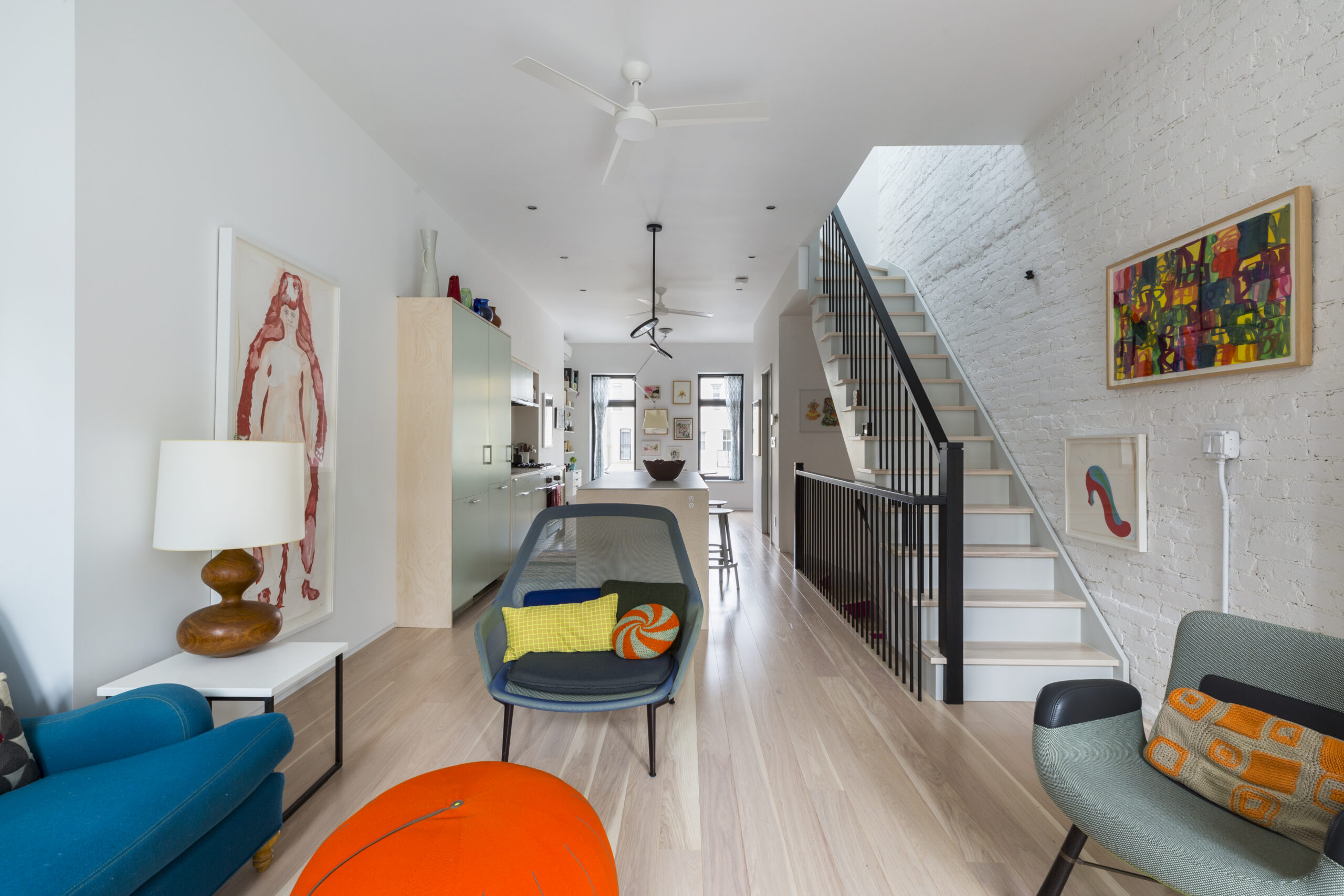 Bronx townhouse remodel with pale wood floors, exposed brick, an open staircase, colorful seating, artwork, and a long sightline toward the kitchen.