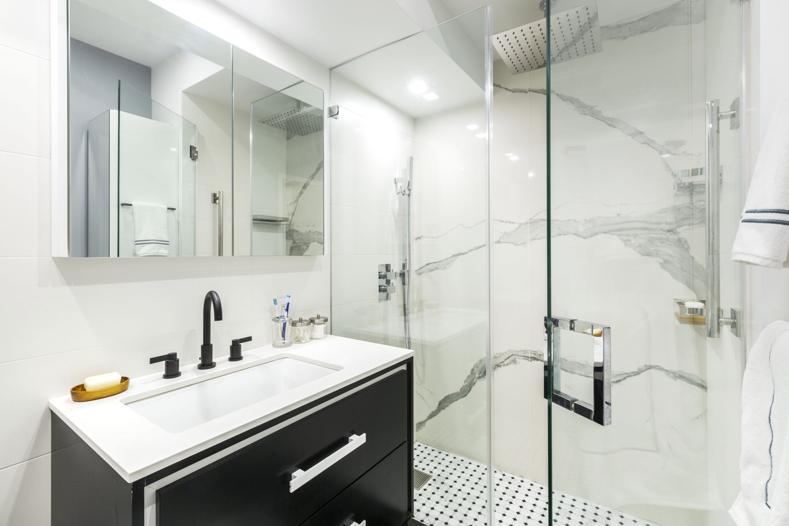 Chelsea bathroom accent wall with Calacatta-look porcelain, black vanity, white countertop, penny tile floor, glass shower doors, and matte black fixtures.