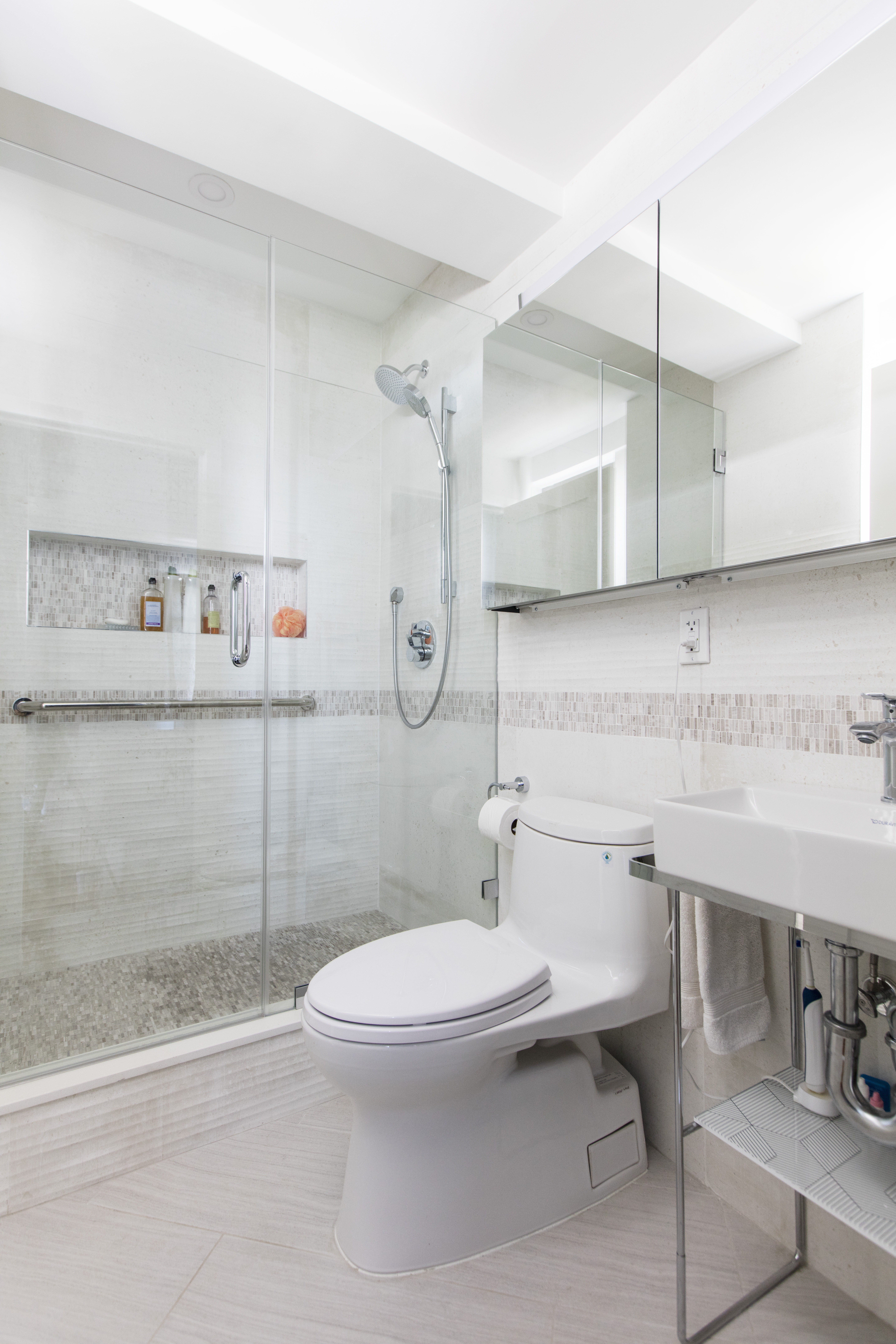 A long shower niche with mosaic tile, grab bars, glass enclosure, and pale stone finishes adds accessible storage to this Kensington bathroom.
