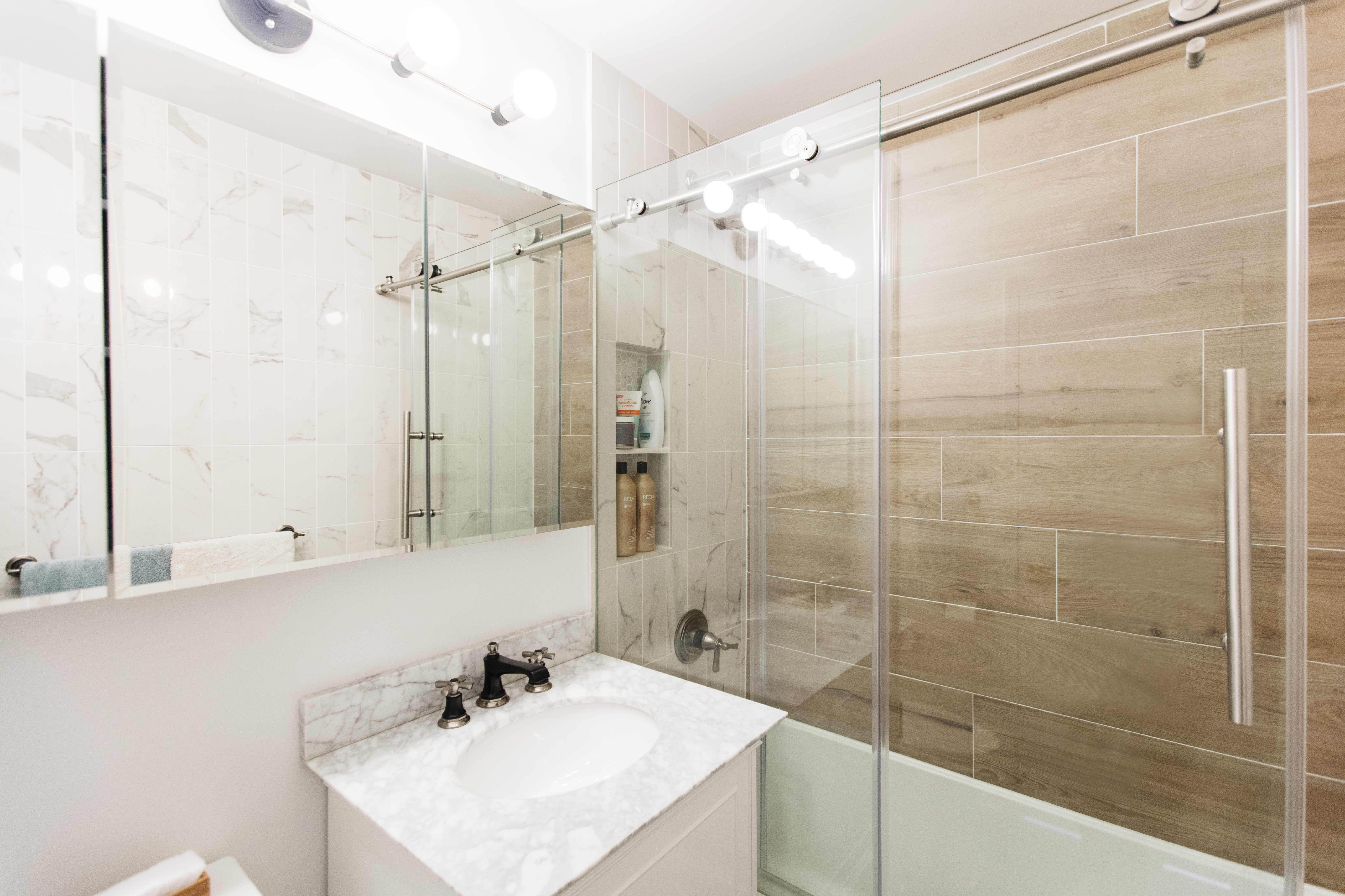 Kips Bay bathroom accent wall with horizontal wood-look shower tile, marble-look wall tile, glass doors, white vanity, and marble countertop.
