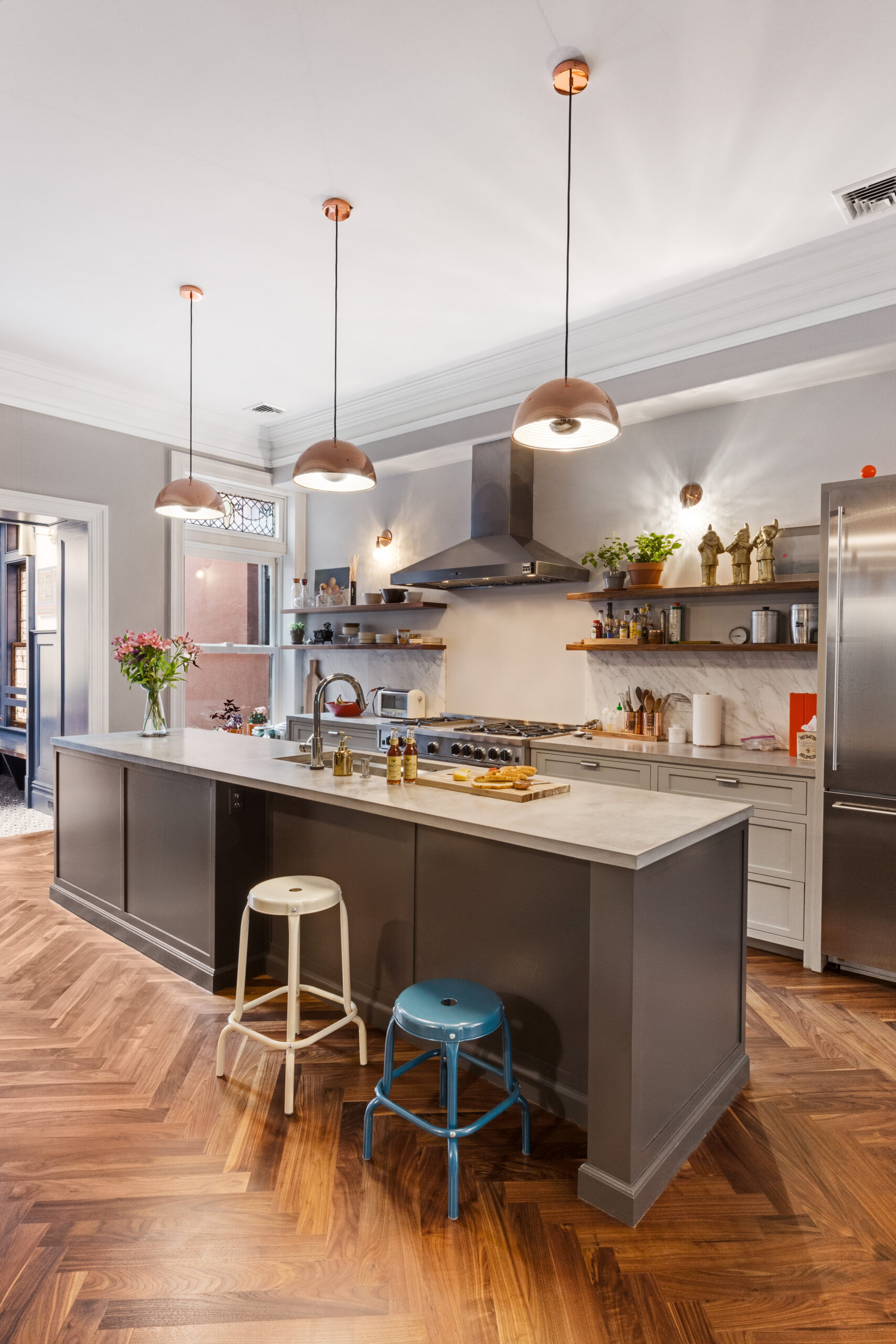 Herringbone walnut flooring, gray island, marble counters, open wood shelves, stainless appliances, and pendant lights in a Brooklyn kitchen.