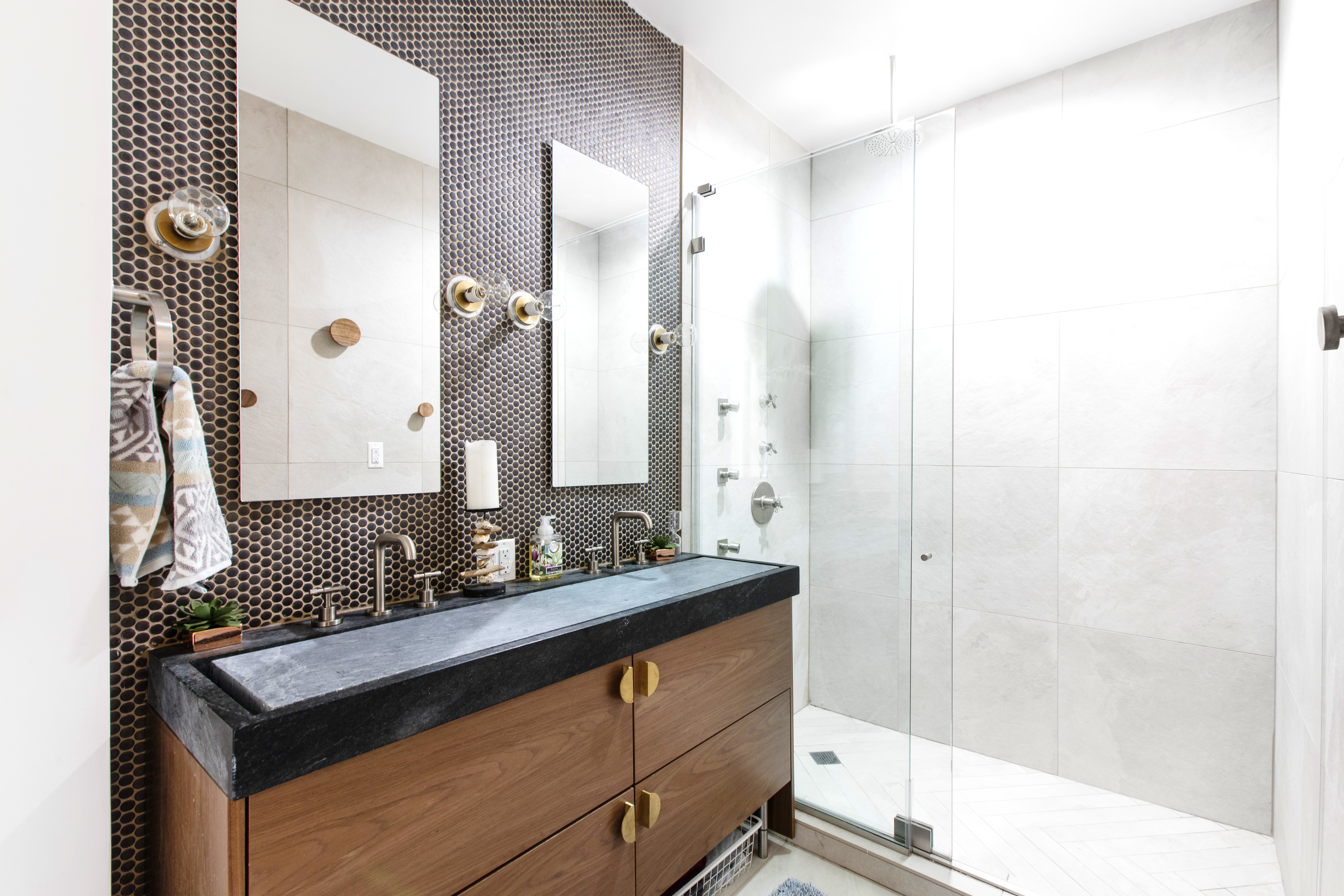 Washington Heights primary bathroom features a custom soapstone double sink, wood vanity, penny tile accent wall, brass hardware, and glass shower.