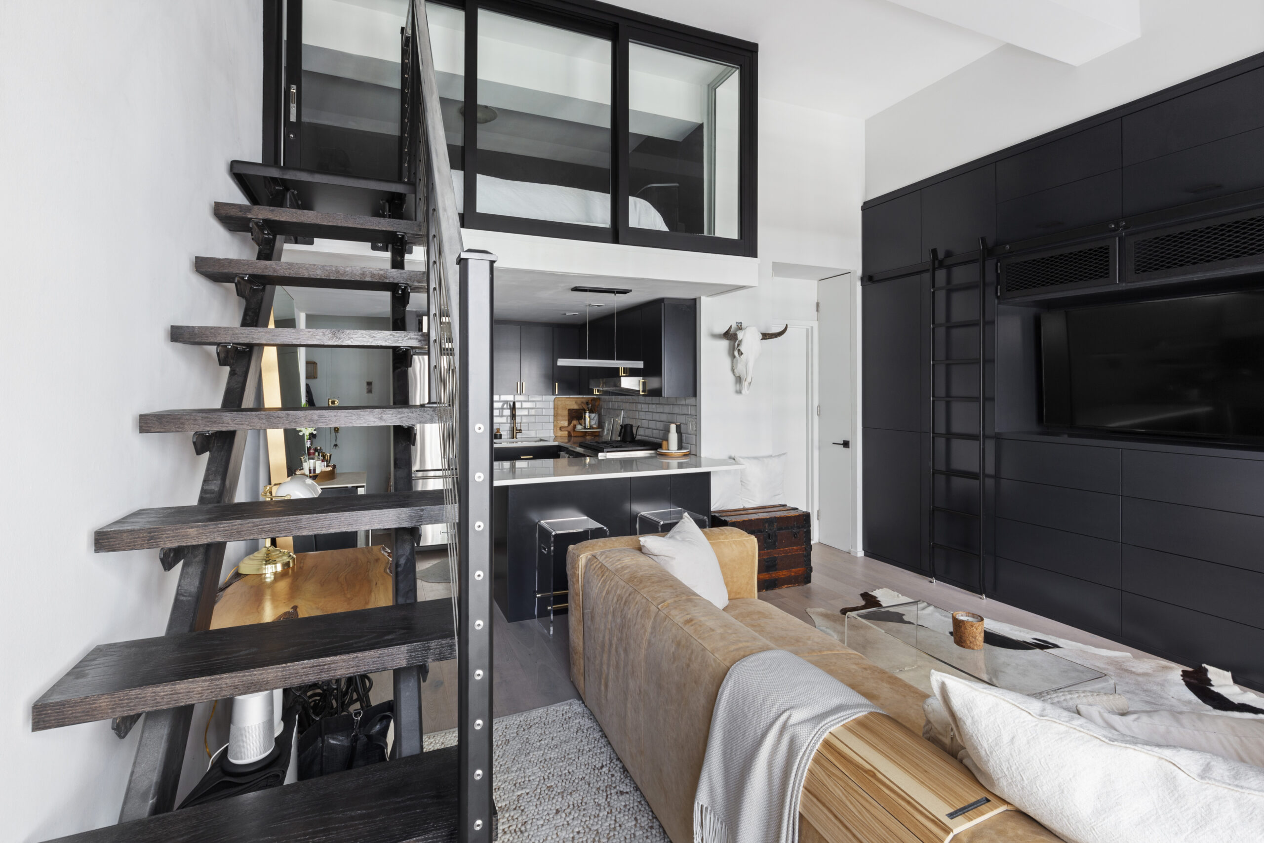 Greenwich Village small apartment with a glass-enclosed sleeping loft, open-riser staircase, black built-in cabinets, and a white kitchen peninsula.