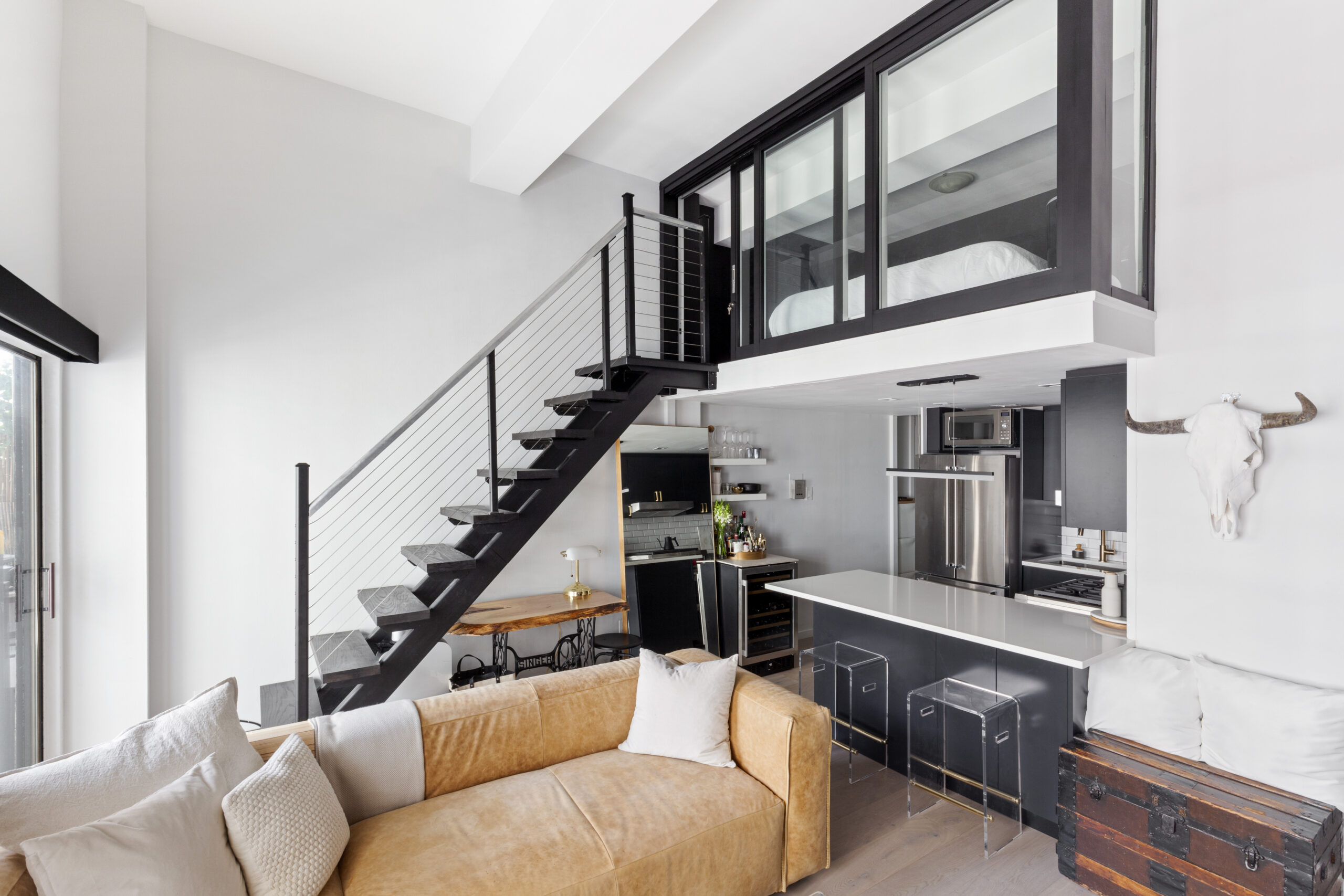 Greenwich Village studio loft with a black metal staircase, glass-enclosed sleeping area, white kitchen peninsula, and an open main living room below.