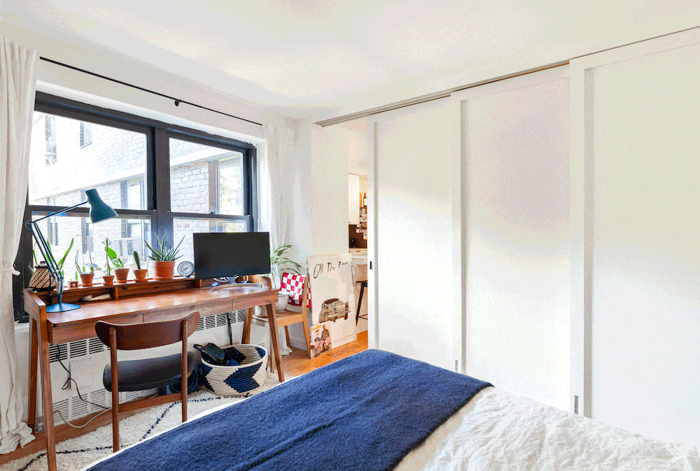Lower East Side small apartment with white sliding doors that separate the bedroom from the living space, plus a wooden desk beside a large window.
