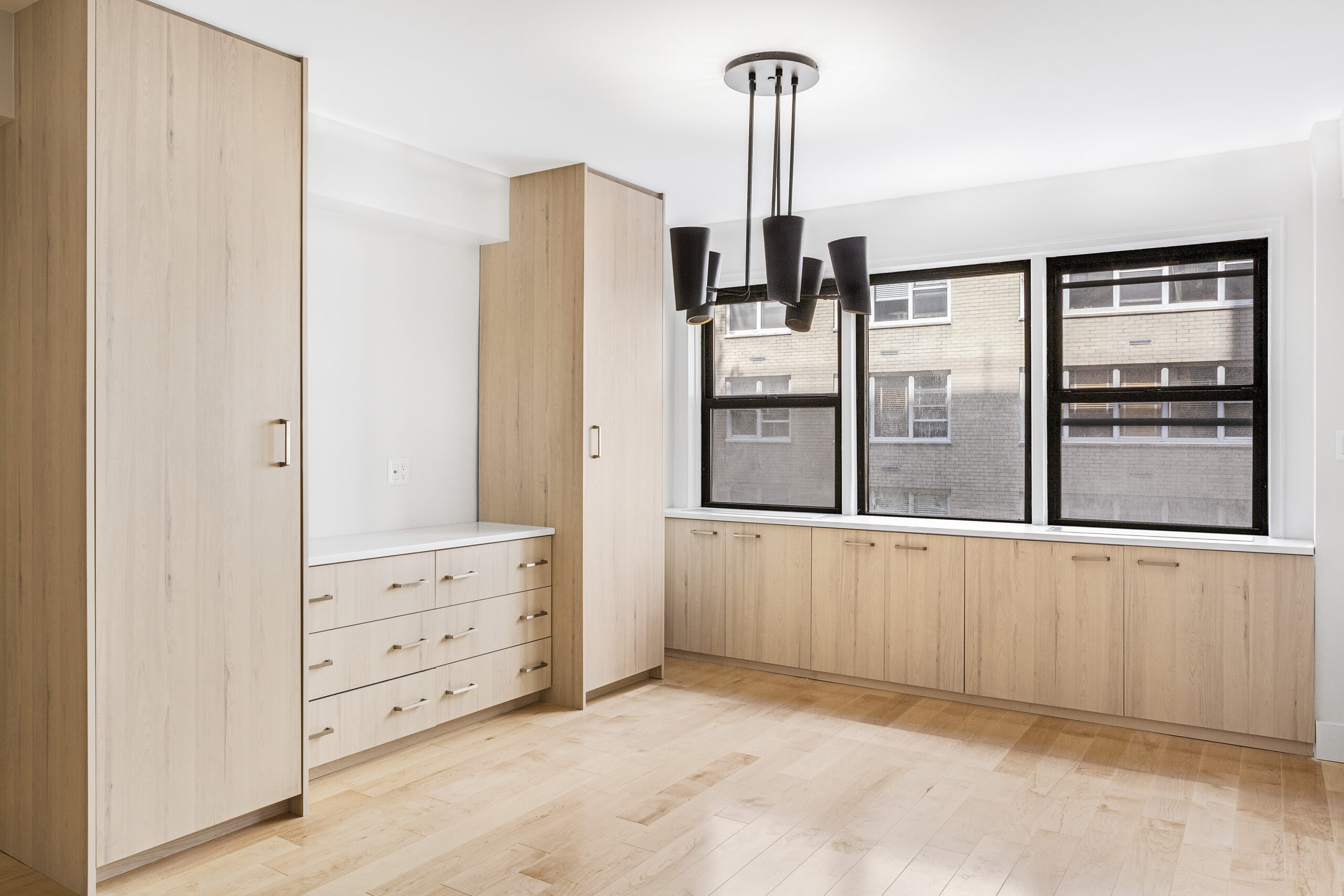 Upper East Side small studio with oak floor-to-ceiling cabinets, built-in drawers below black-framed windows, light maple flooring, and a chandelier.