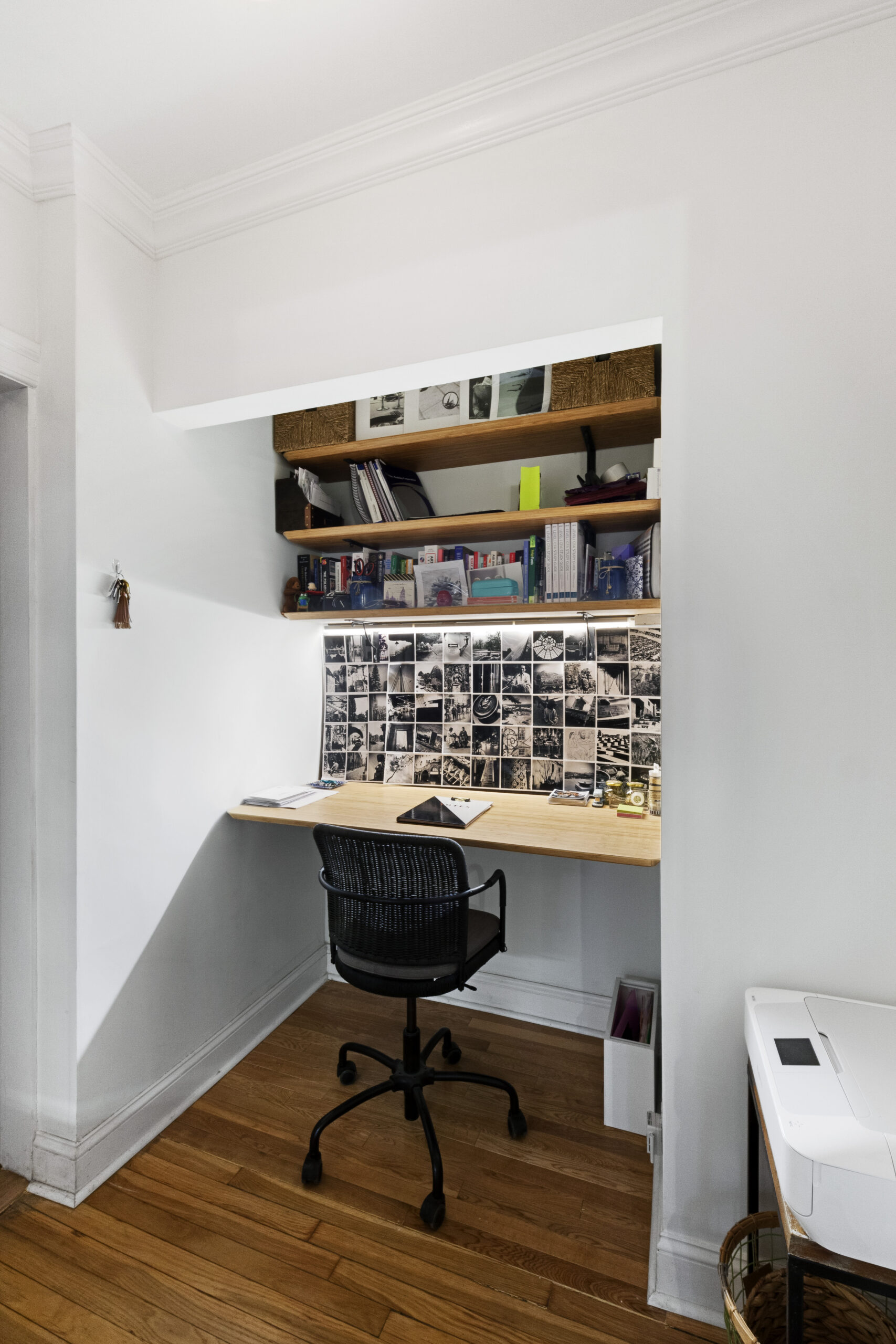 Upper West Side studio apartment office nook with a built-in wood desk, floating shelves, photo display, black swivel chair, and refinished wood floors.